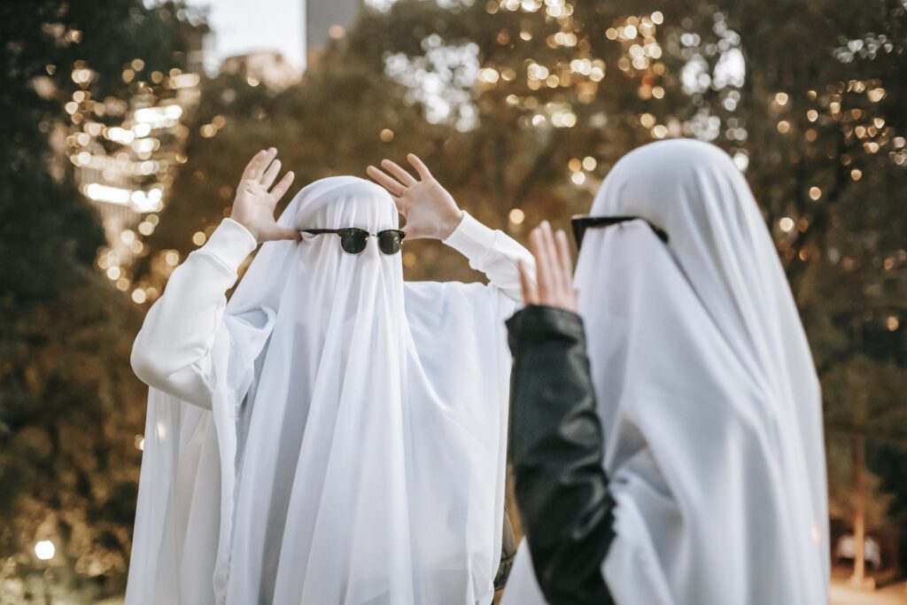 Cheerful anonymous couple with raised arms wearing ghost costumes and sunglasses looking at each other and having fun while standing in park with trees on blurred background during Halloween