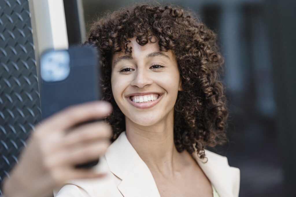 Positive young ethnic female with curly brown hair taking self shot on cellphone in town on blurred background